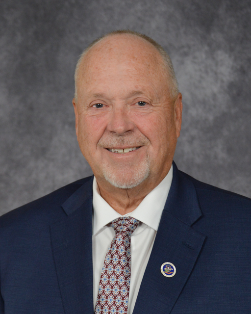 male wearing a blue blazer, white collared shirt with a red and blue patterned neck tie. State of north dakota pinned on right collar.
