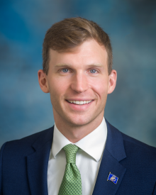 male wearing a navy suit jacket with a lime green polka dotted neck tie and a north dakota emblem pin on his chest.